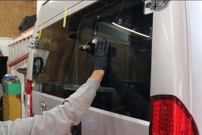 Gloved worker applies a tool to the rear window of a white van in a workshop.