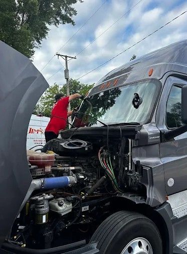 Person in red shirt working under the open hood of a gray truck outdoors.