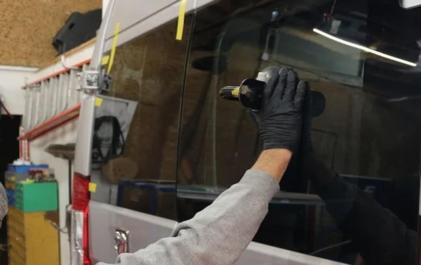 Gloved worker using a drill on a glossy black vehicle panel in a workshop.
