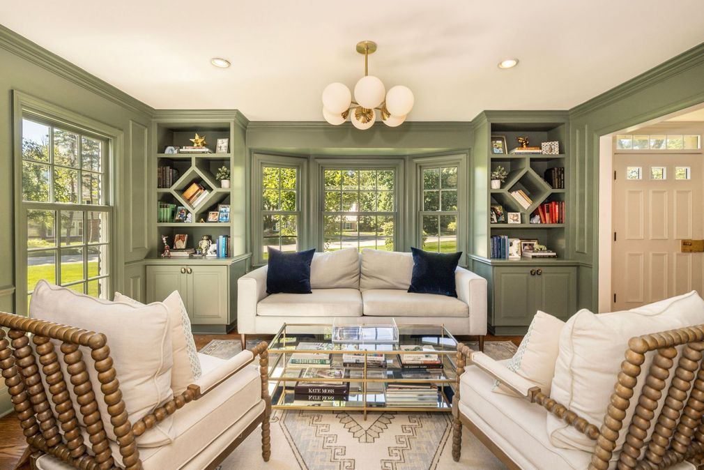 A light-filled living room features sage green built-in shelves flanking a sofa, two beaded armchairs, and a glass table.