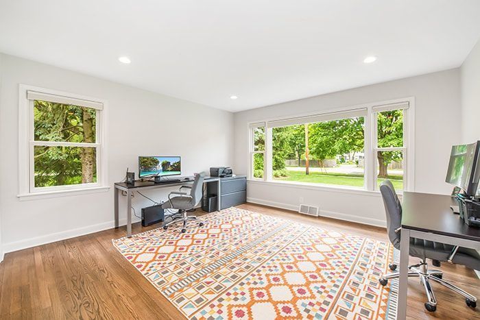 Home office with two desks, a patterned rug, and large windows overlooking a yard.