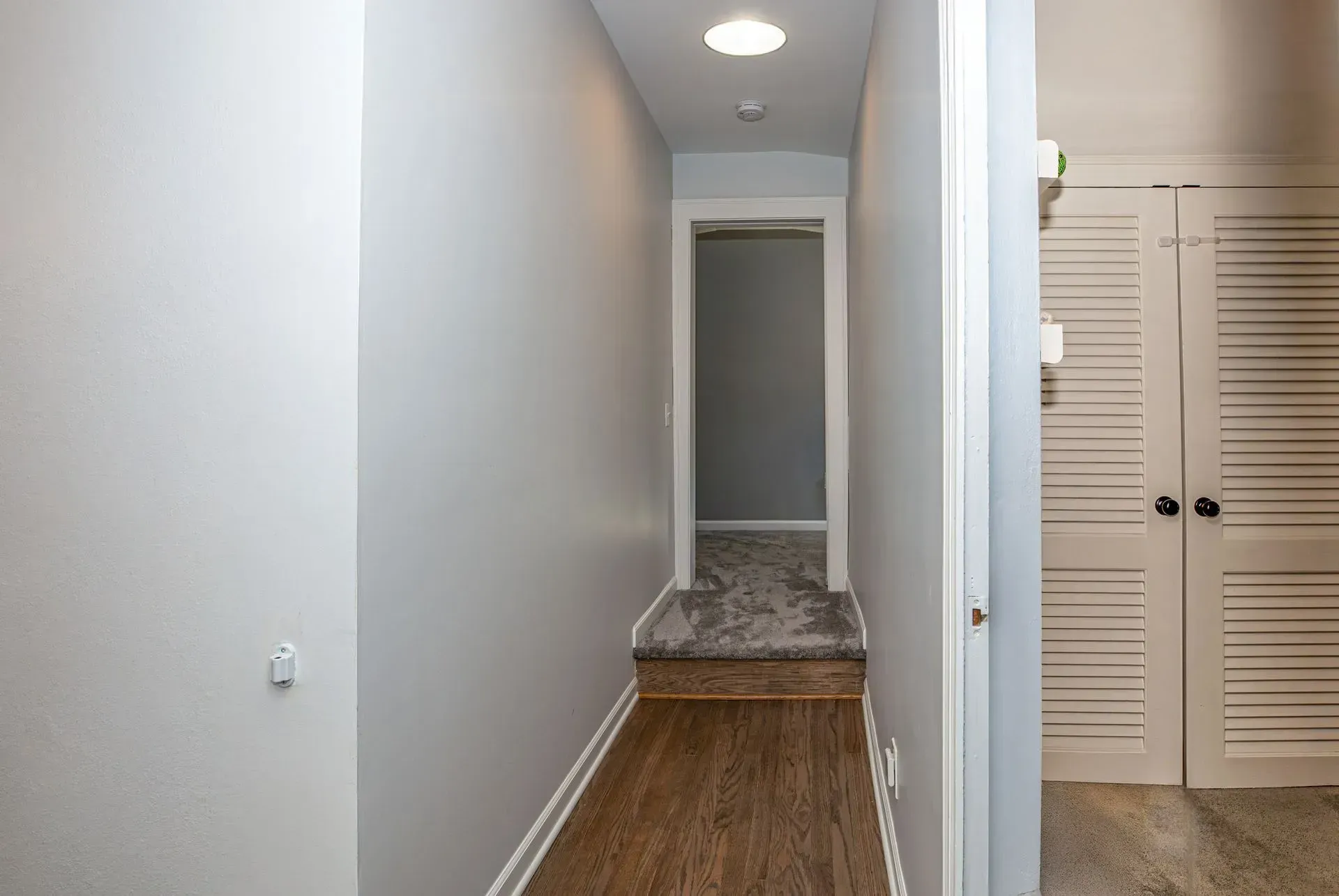 Narrow hallway with gray walls, wood-look floor, carpeted steps, and a doorway. A closet is visible on the right.