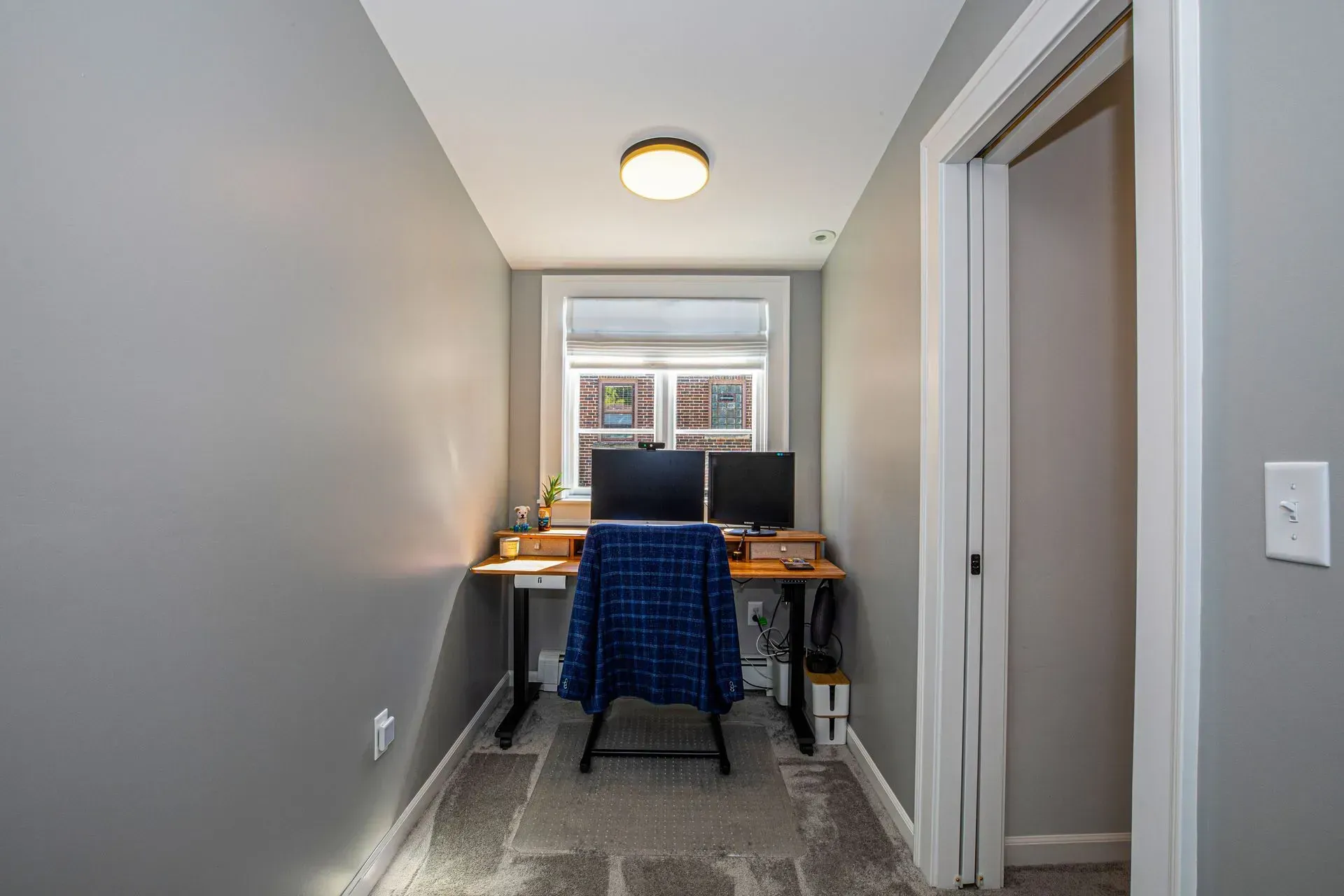 Narrow home office with desk, two monitors, and window; gray walls, carpet, and a closed door.
