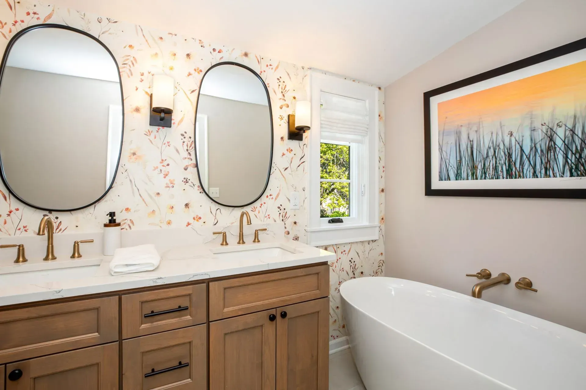 Bathroom with double vanity, oval mirrors, floral wallpaper, and a freestanding tub.