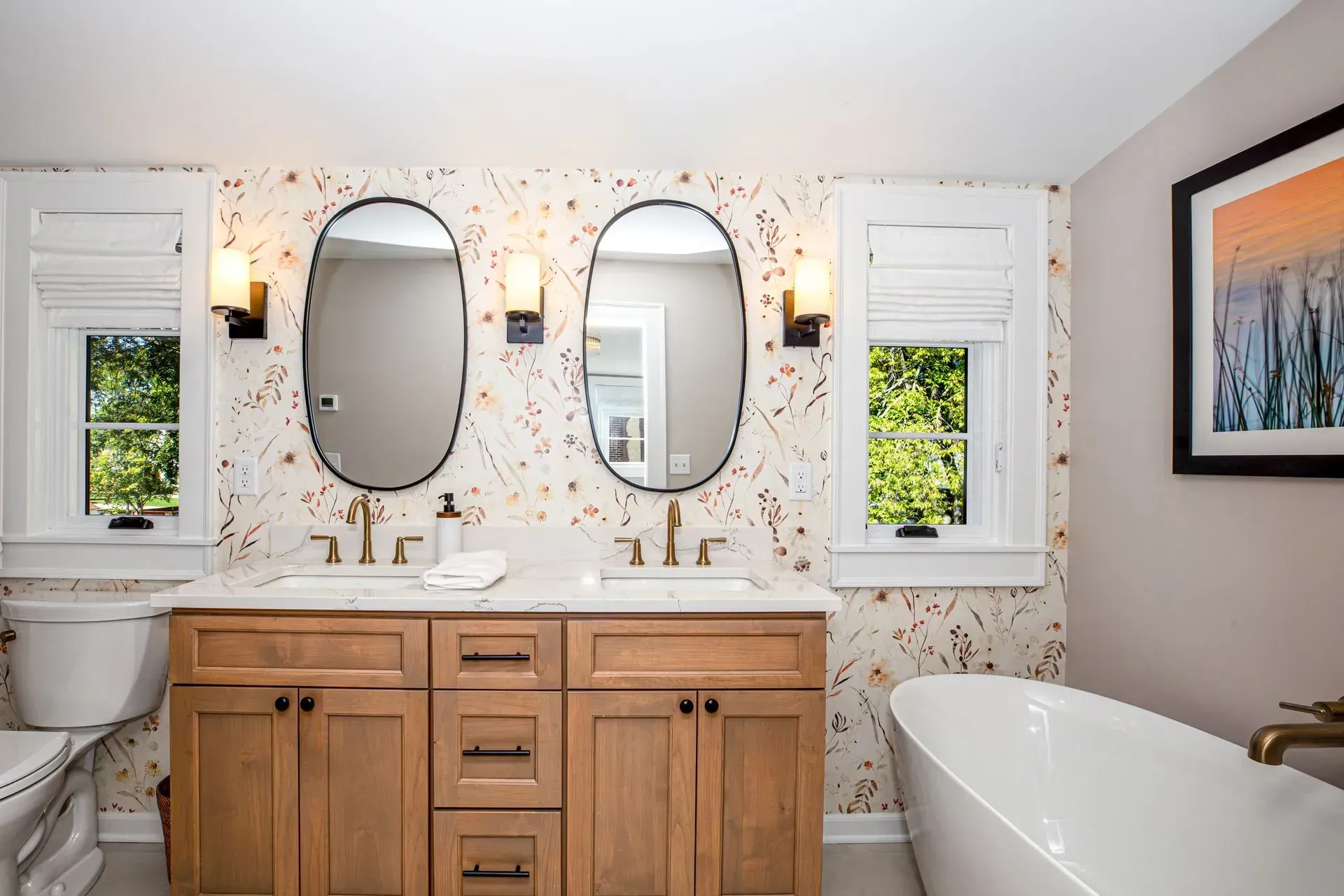 Bathroom with double vanity, oval mirrors, floral wallpaper, and a bathtub.