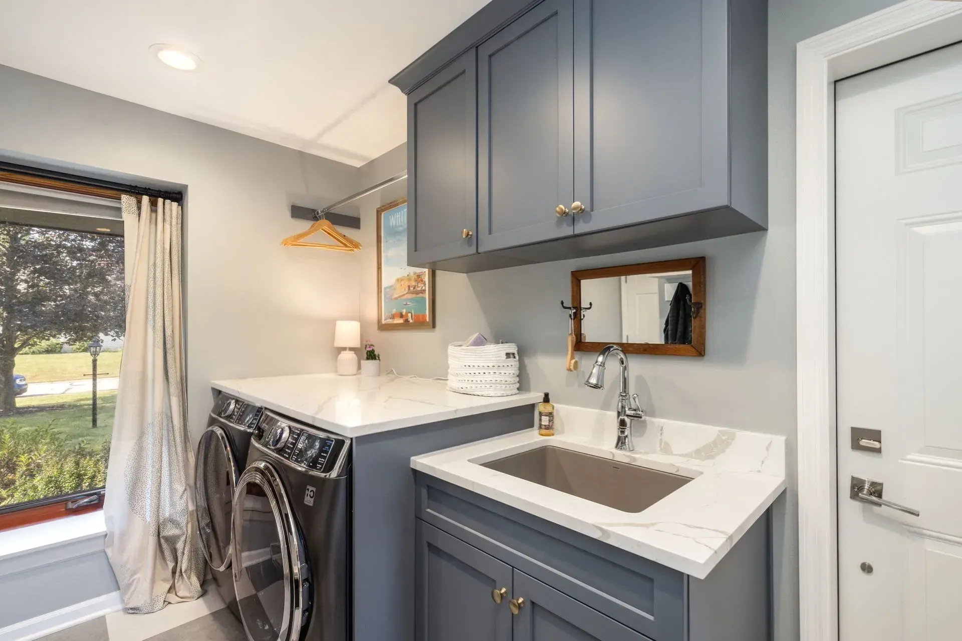 Laundry room with blue cabinets, washer/dryer, and a sink. White countertop and a window with a view.