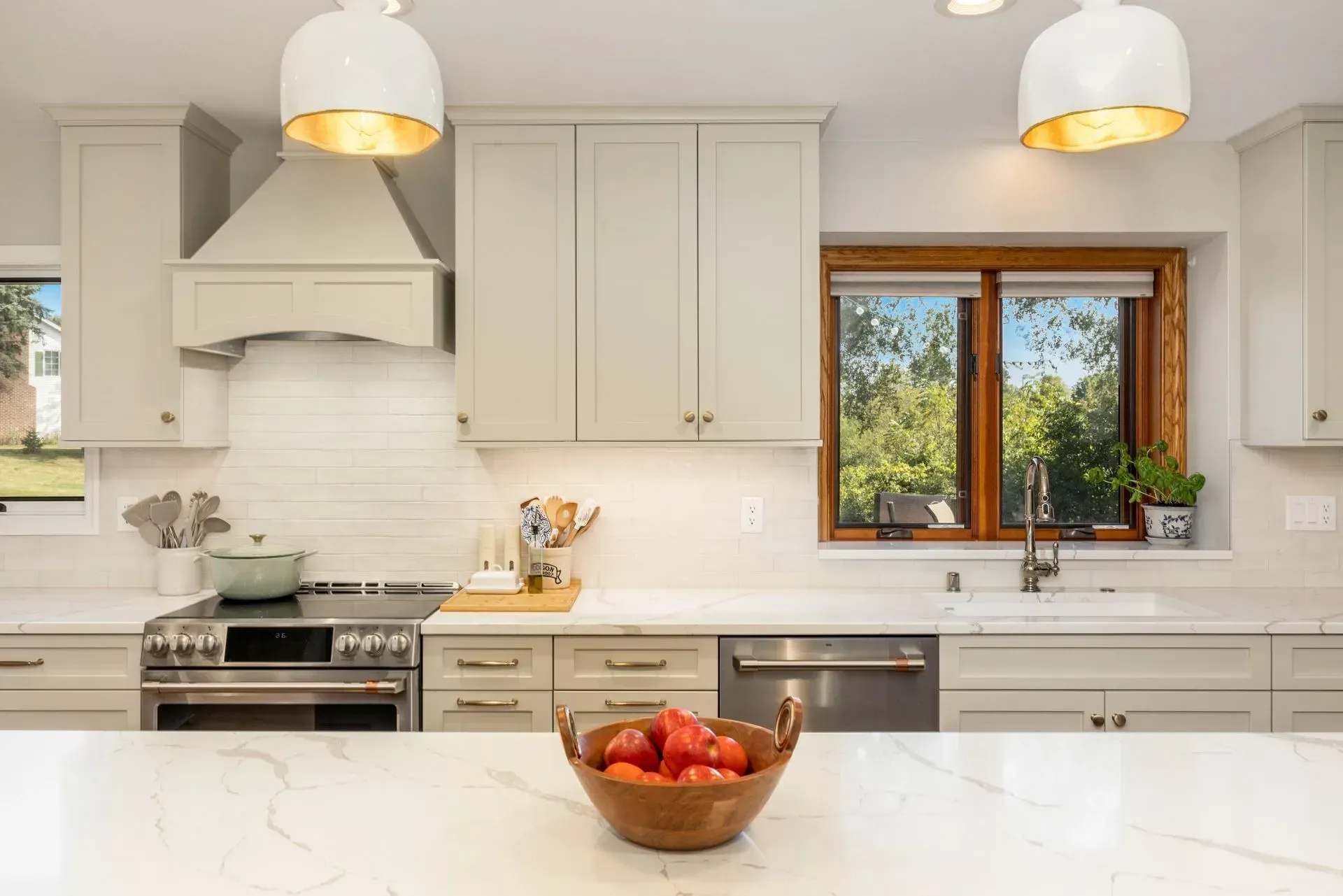 Modern kitchen with light grey cabinets, stainless steel appliances, and a wooden bowl of tomatoes on countertop.