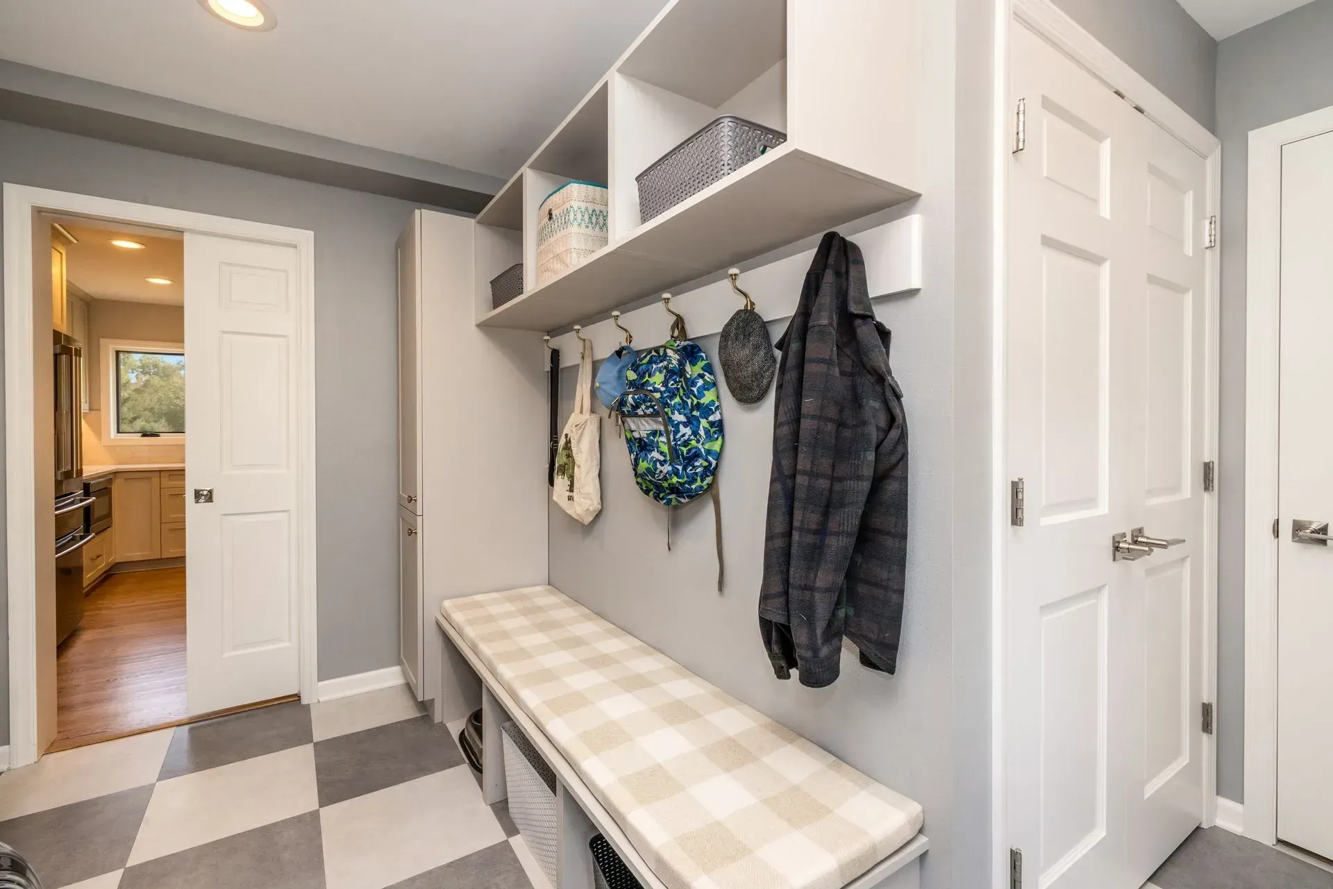 Mudroom with bench, hooks, storage, and a door to a kitchen, grey and white checkered floor.