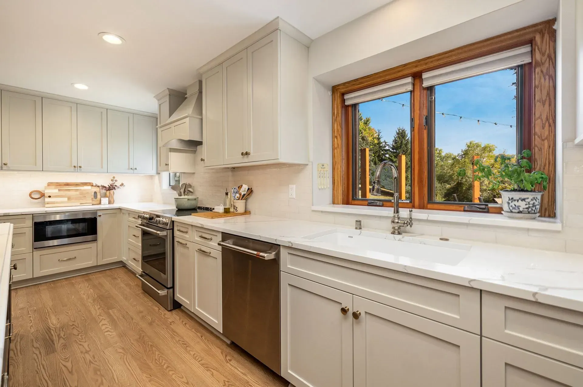 Modern kitchen with light gray cabinets, stainless steel appliances, wooden window frame, and wood flooring.