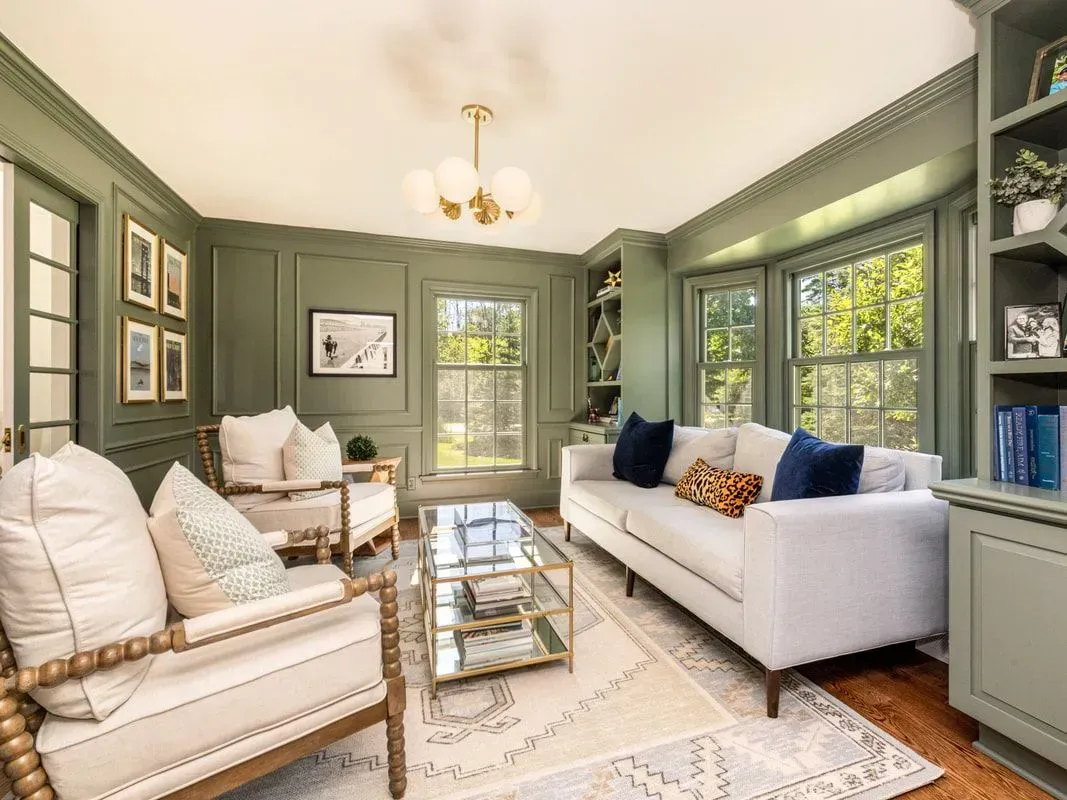 A cozy living room with olive green walls, white furniture, and a glass coffee table.