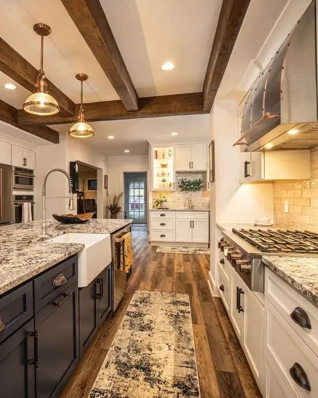 Kitchen with dark blue island, white cabinets, granite countertops, wood beams, and copper pendant lights.