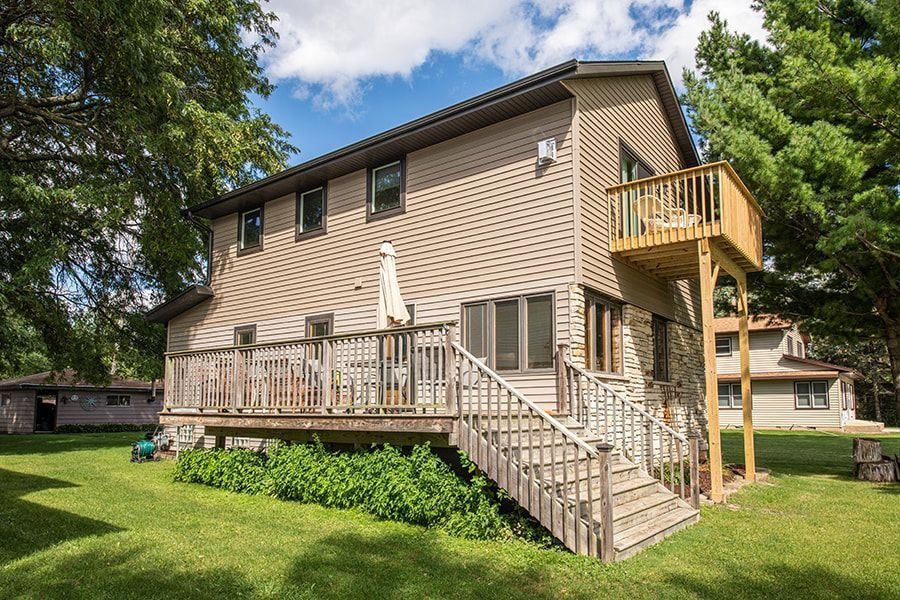 Two-story house with a deck and small balcony. Green lawn and trees surround it.