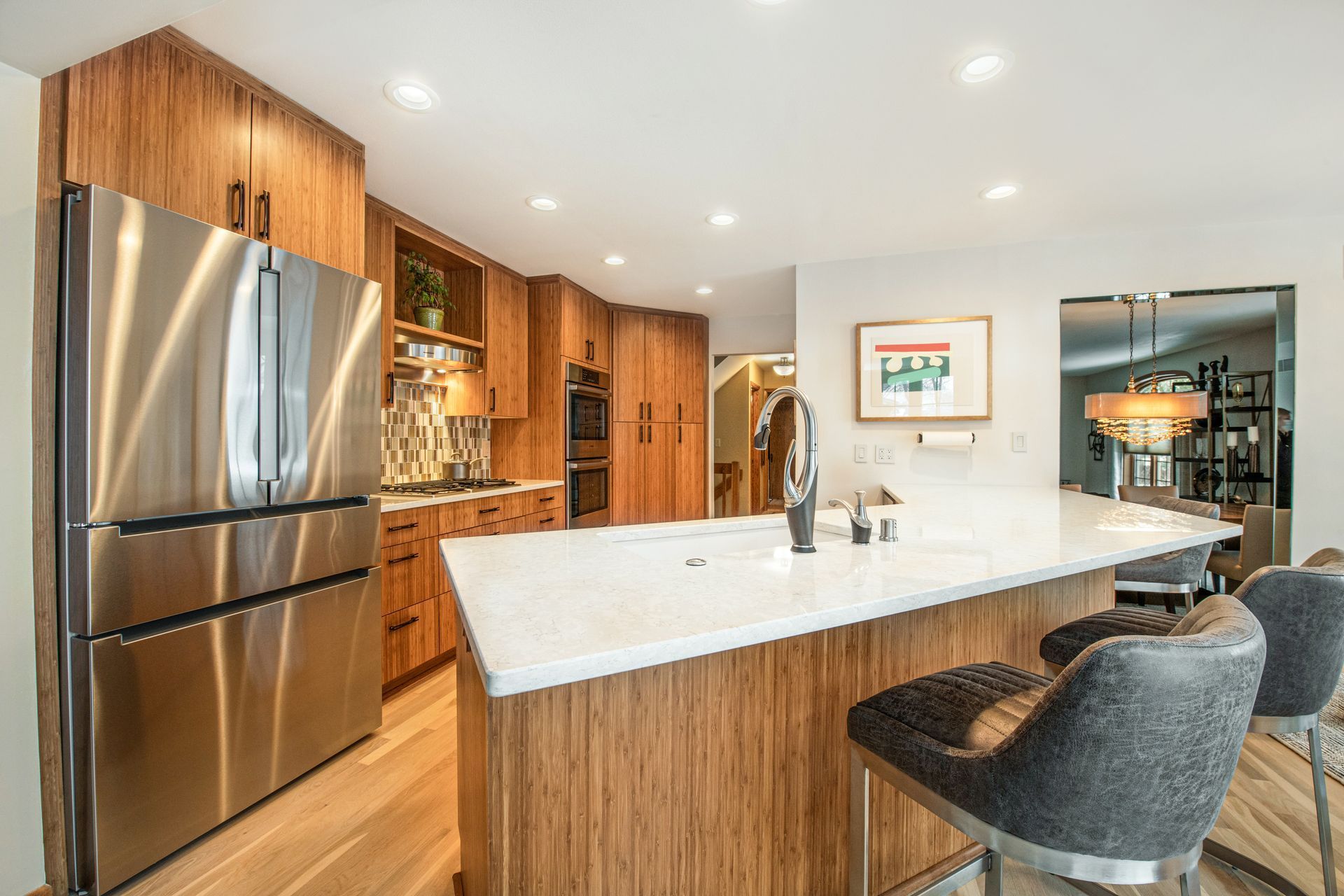 Modern kitchen with wood cabinetry, stainless steel refrigerator, white island countertop, and gray bar stools.