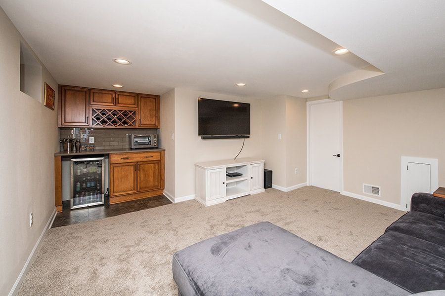 Basement with a wet bar, TV, and gray carpet. The room has a white cabinet for storage and a black couch.
