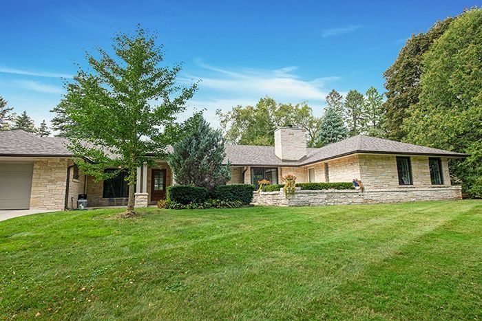 Single-story stone home with a grassy yard, trees, and blue sky.