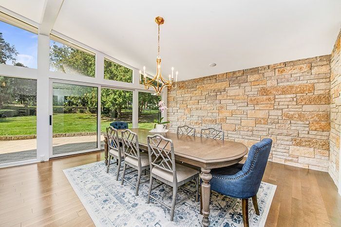 Dining room with stone wall, oval table, chairs, rug, and large windows overlooking a yard.