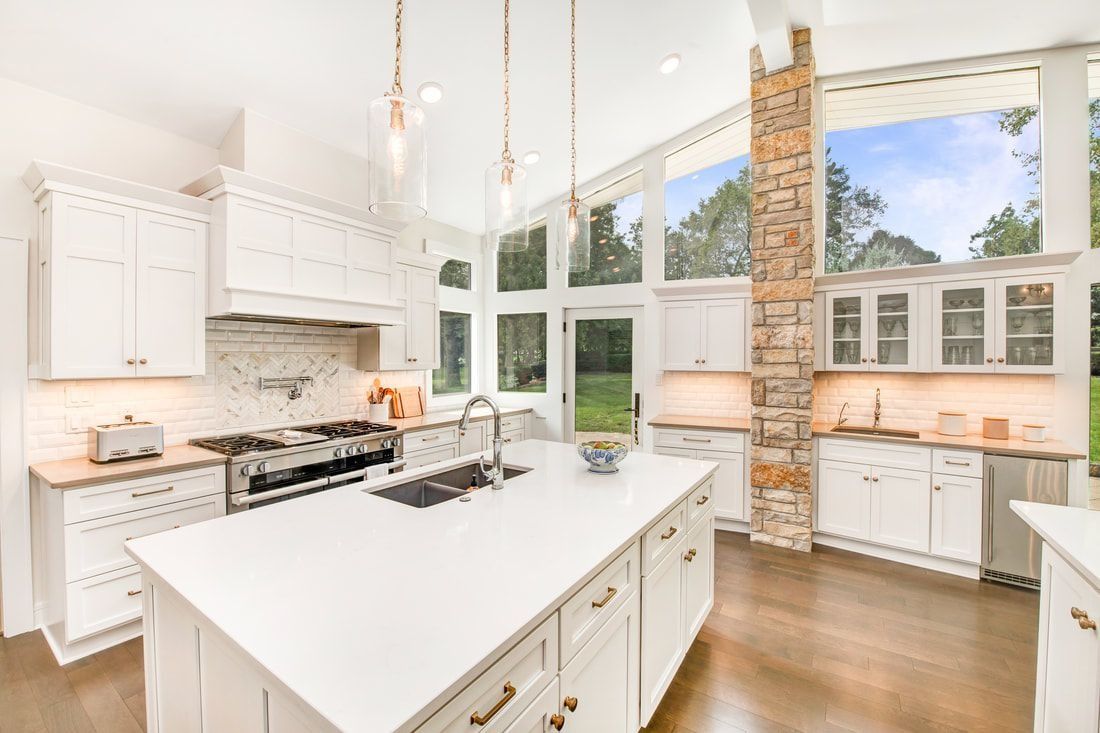 Bright white kitchen with large island, tall windows, stone accent, and wooden floors.