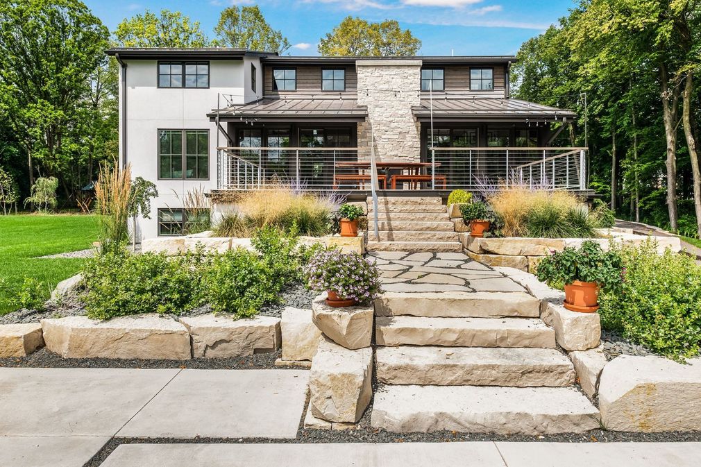 Modern two-story home with stone steps, patio, and landscaping. Green lawn and trees surround the house on a sunny day.