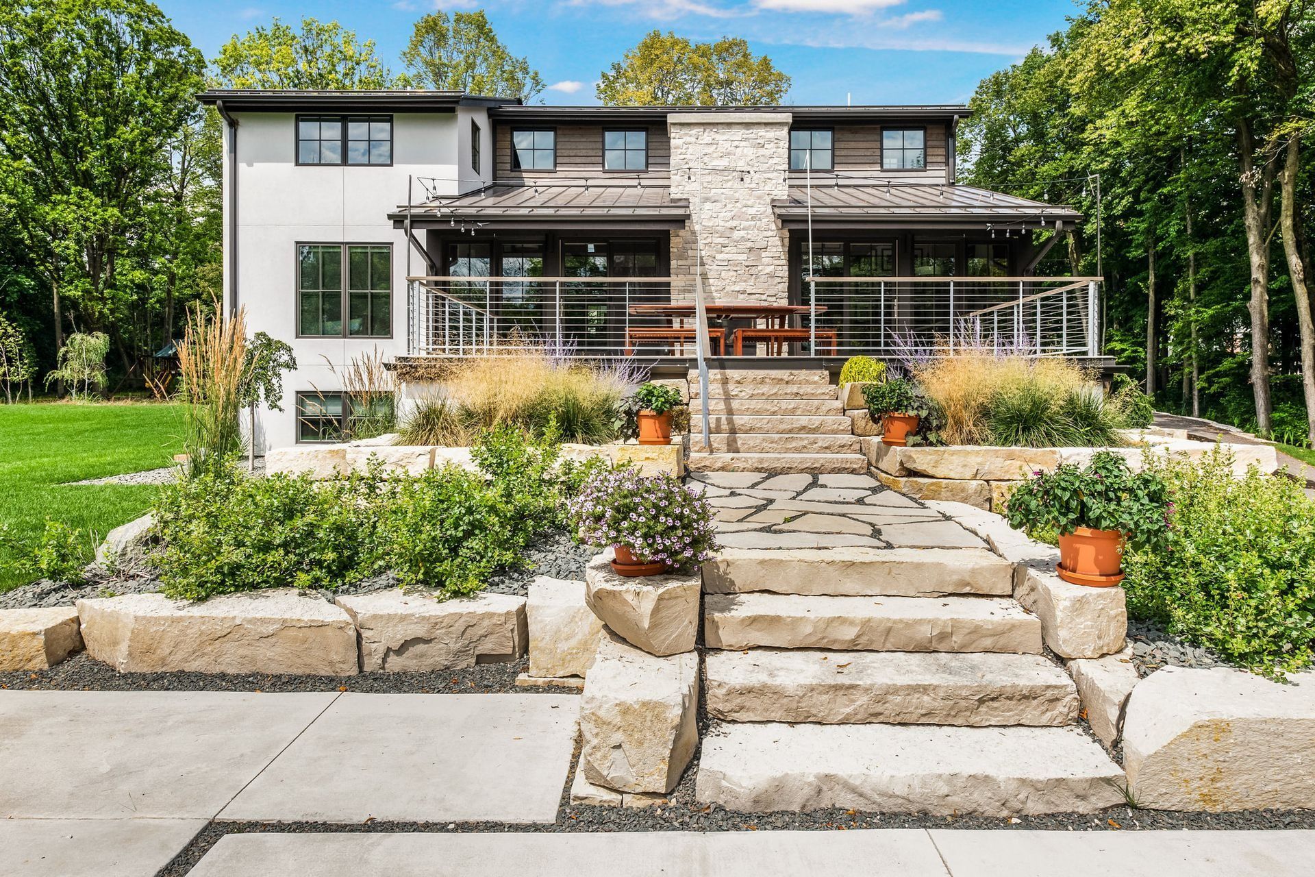 Modern two-story home with stone steps, patio, and landscaping. Green lawn and trees surround the house on a sunny day.