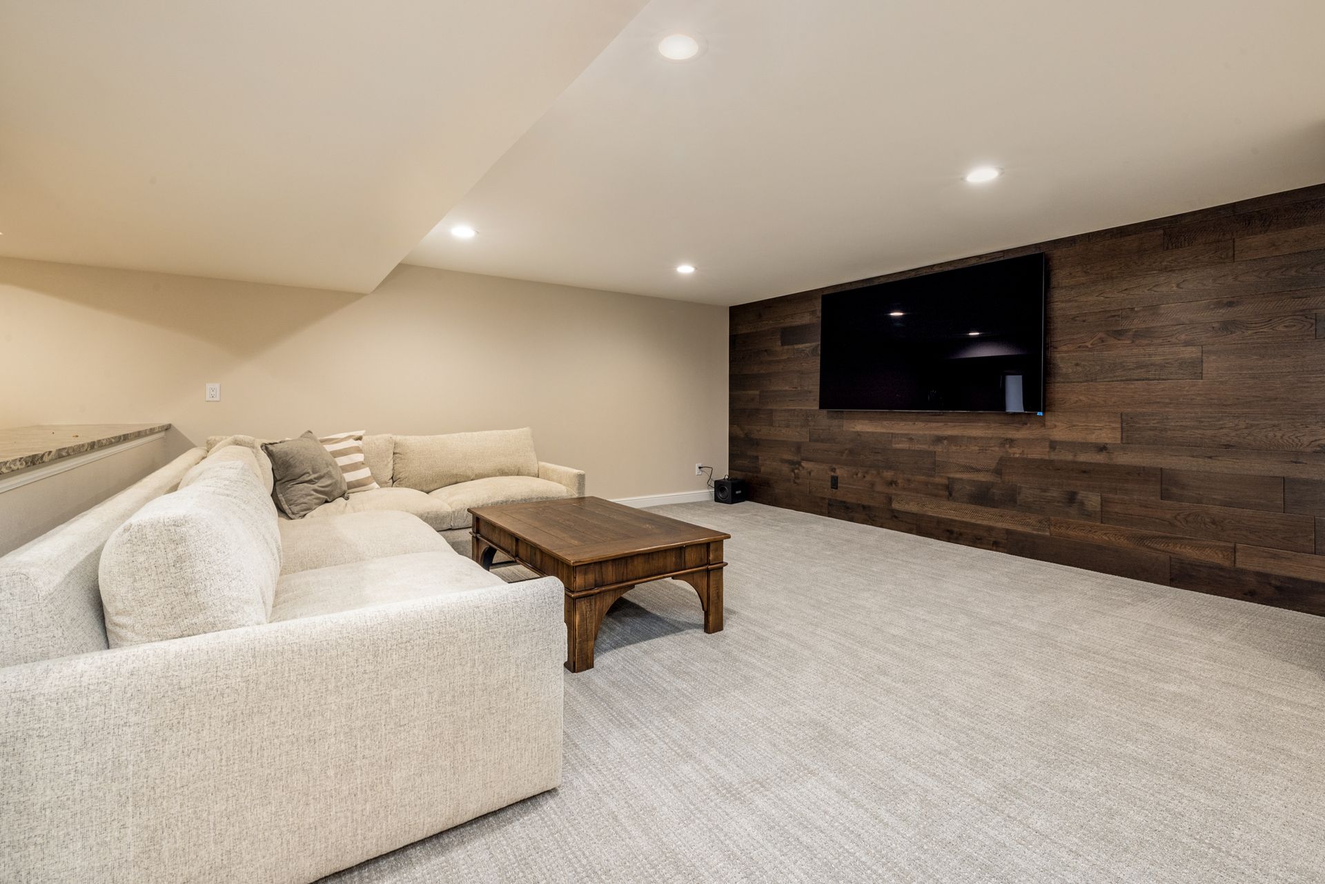 Cozy basement living room with a large TV mounted on a wood-paneled wall, light-colored sectional sofa, and a coffee table.