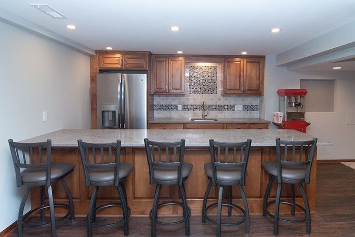 Bar with granite countertop and brown cabinets, stainless steel fridge, and six barstools.
