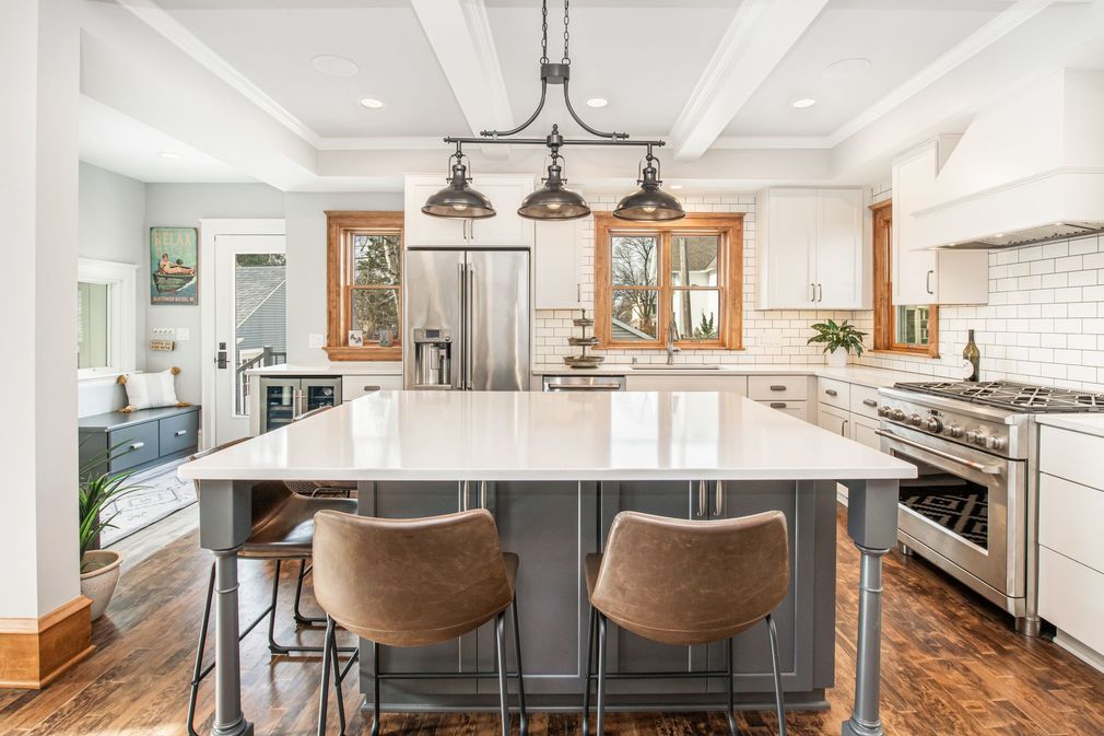 A modern kitchen features a large grey island with stools under a three-light pendant fixture, white cabinets, and wood floors.