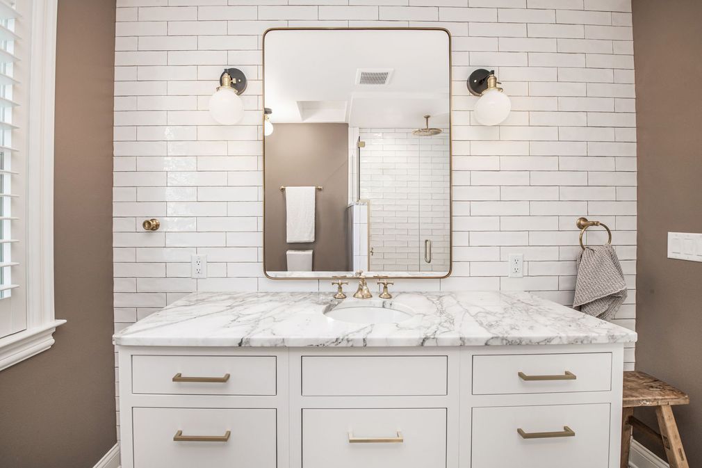 Bright bathroom vanity with marble countertop, gold fixtures, and a large framed mirror against white tiled walls