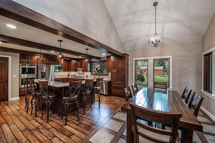 Spacious kitchen with island, dining table. Wooden cabinets, beams, and floors; light gray walls, windows with backyard view.