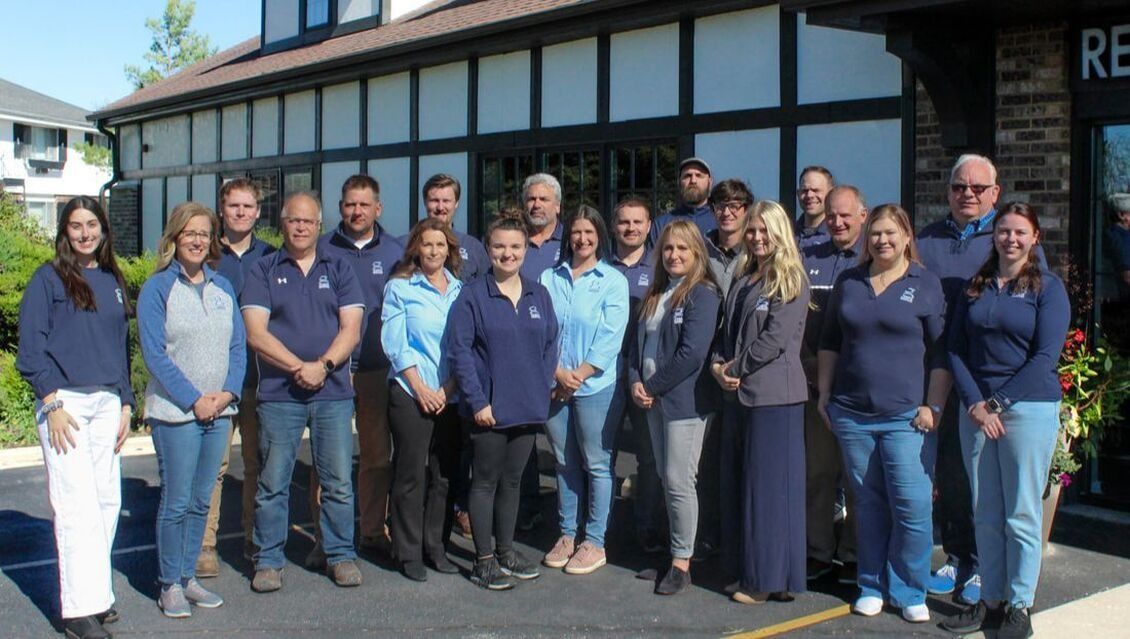 Group of people posing in front of a building, many wearing matching blue shirts.