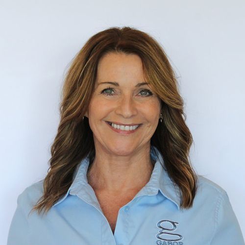 Linda with wavy brown hair smiling, wearing a light blue collared shirt with logo against a white background.