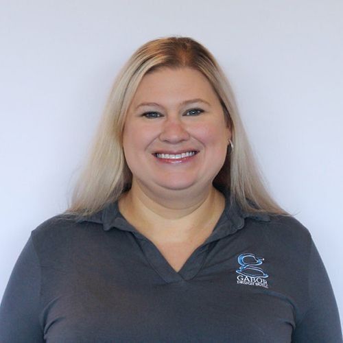 Tara with blonde hair smiles, wearing a gray polo shirt with a logo, against a white background.