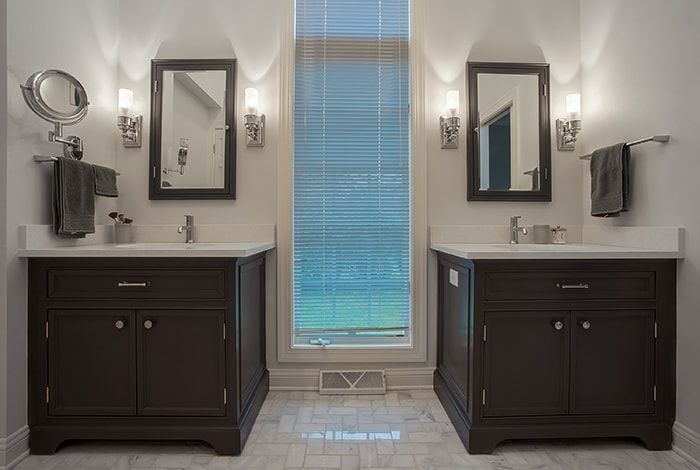 Two dark brown bathroom vanities with mirrors and sconces flank a window; white countertops.