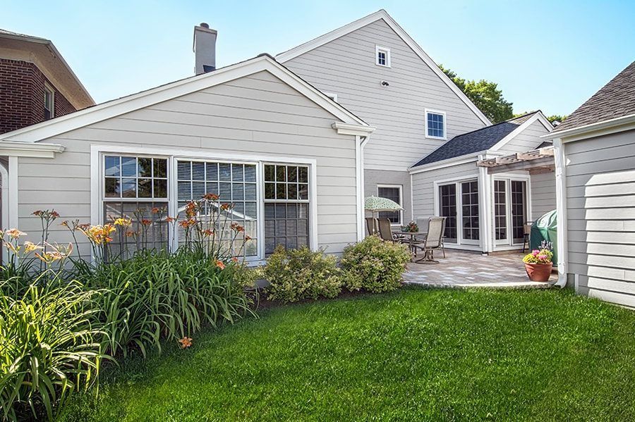 Backyard with gray house, patio, green grass, and plants.