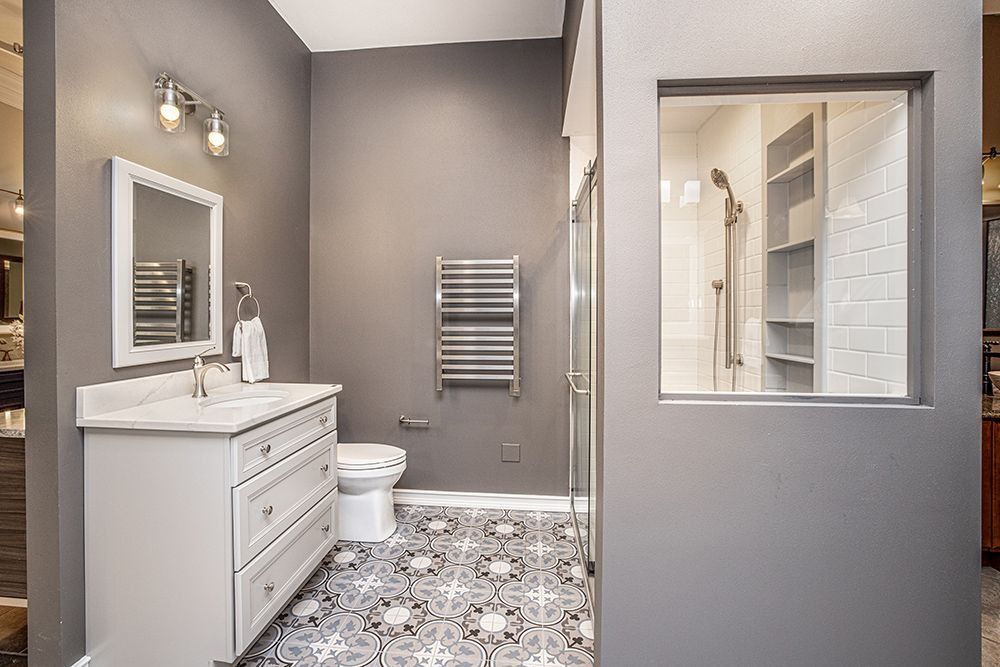 Bathroom with gray walls, white vanity, patterned floor, and shower seen through a window.