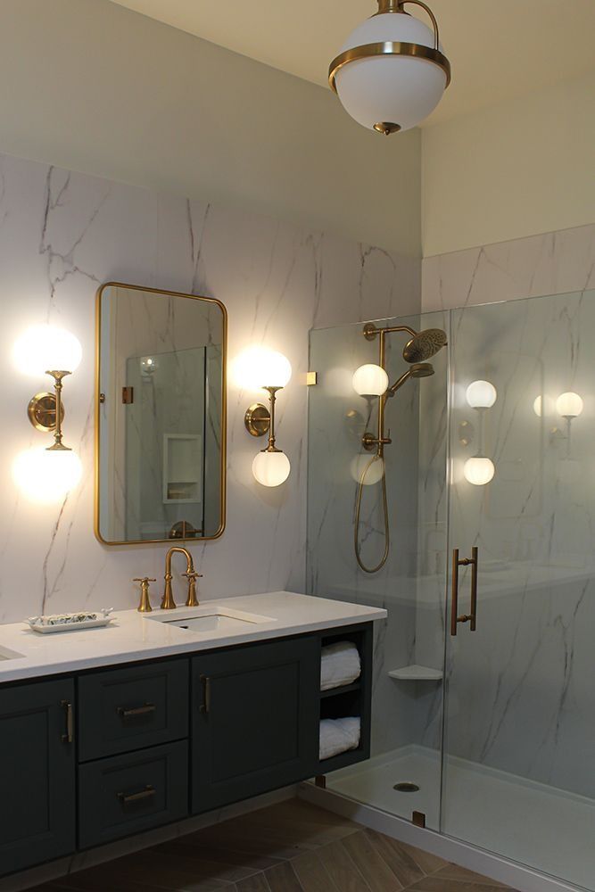 Bathroom with vanity, mirror, shower, and globe lights. Features gray-green cabinet, white countertop, marble-look walls, and gold fixtures.