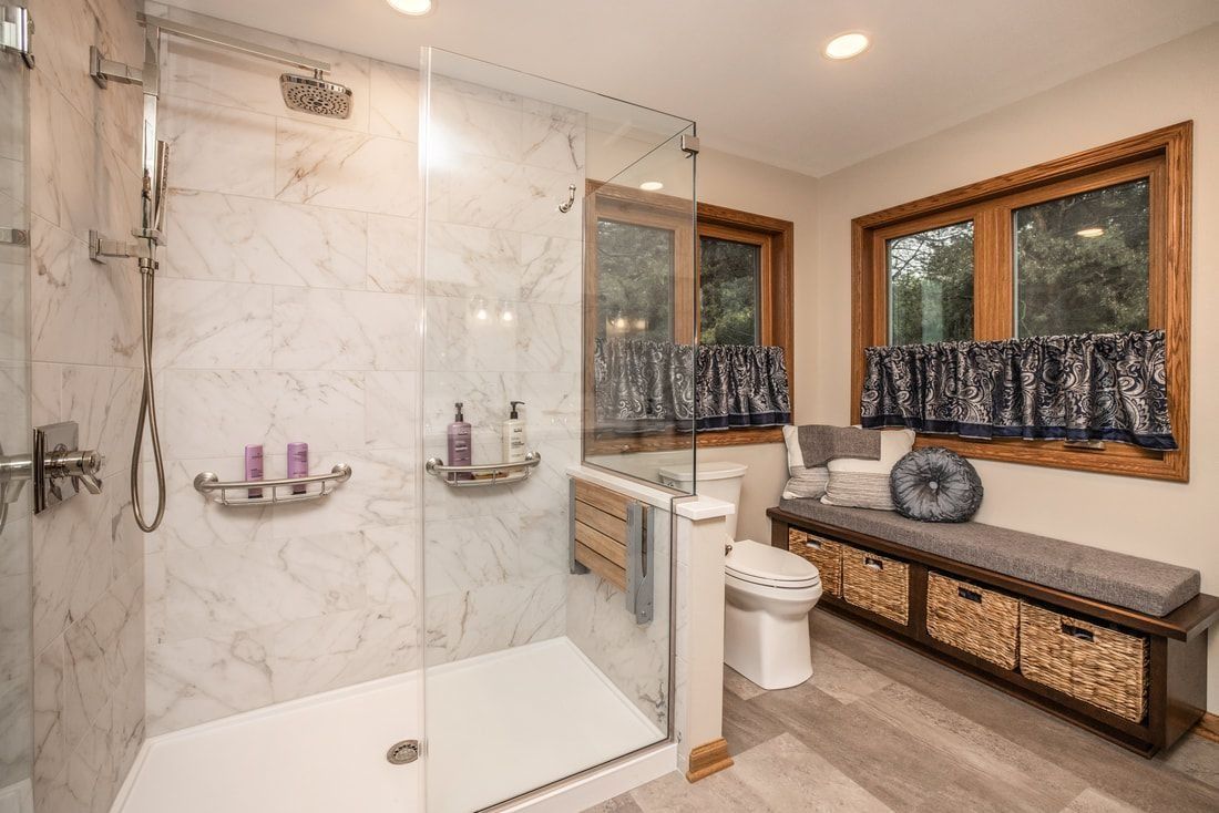 A modern bathroom with a walk-in shower, bench, and two windows; white, marble, and wood accents.