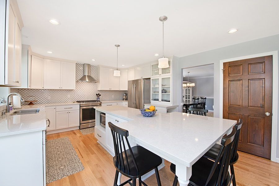 Modern white kitchen with island, hardwood floors, stainless steel appliances, and a dark wooden door.