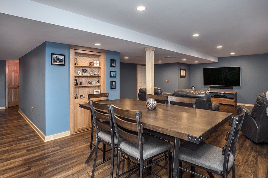 Blue-walled basement with a dining table, built-in shelves, and a TV. Wooden floor, gray chairs.