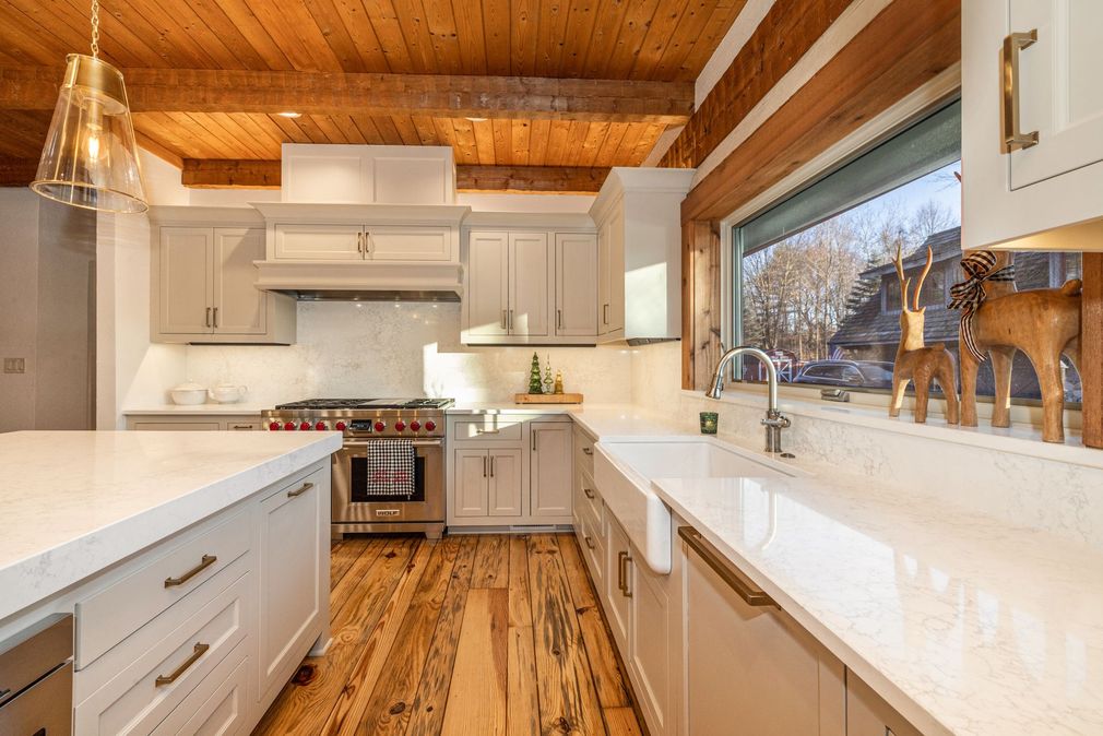 A modern, white farmhouse kitchen with wooden ceiling beams, a large island, and a sink beneath a window with deer decor.