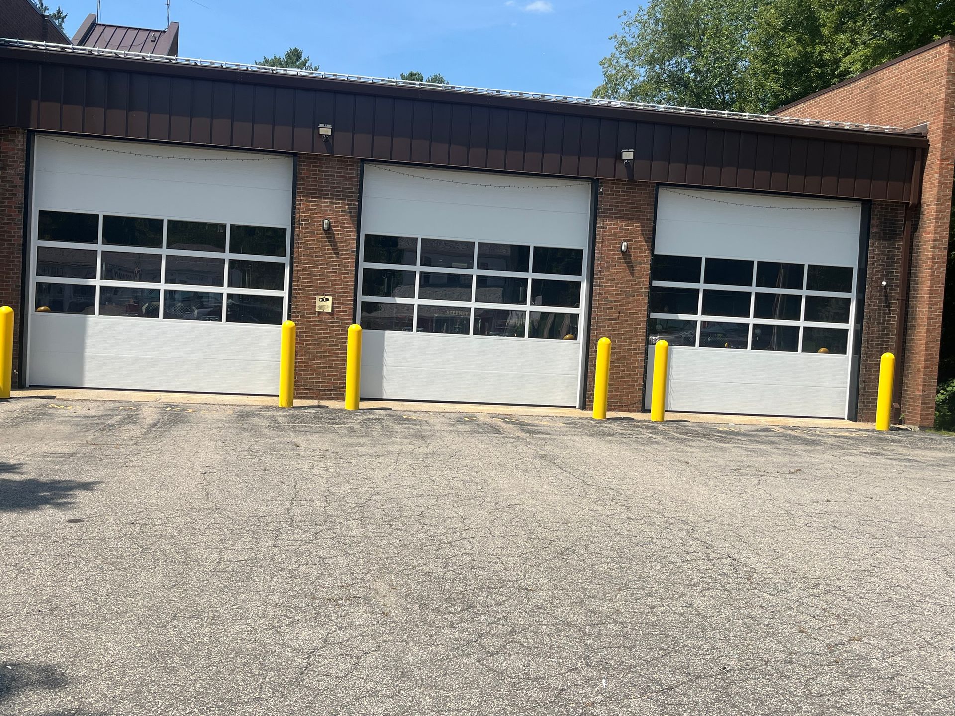 Three white garage doors with glass panels, flanked by yellow poles, on a brick building.