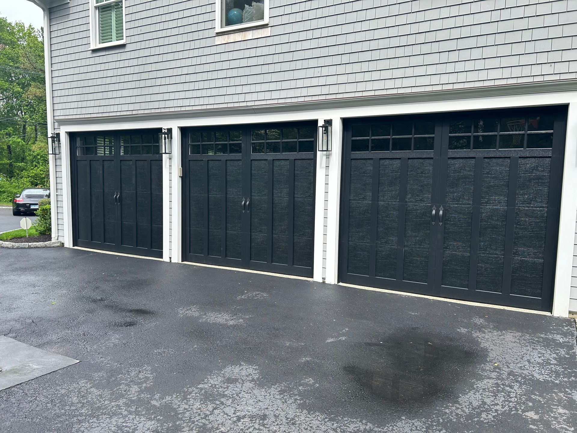 Three black garage doors with windows on a gray house, asphalt driveway.