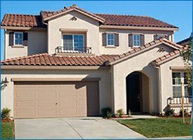 Two-story beige house with red tile roof, brown garage door, and arched front entrance.