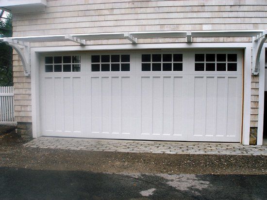 White garage door with window panels, under a pergola, in front of a shingled house.