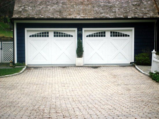Two white garage doors with decorative windows, on a blue building with a brick driveway.
