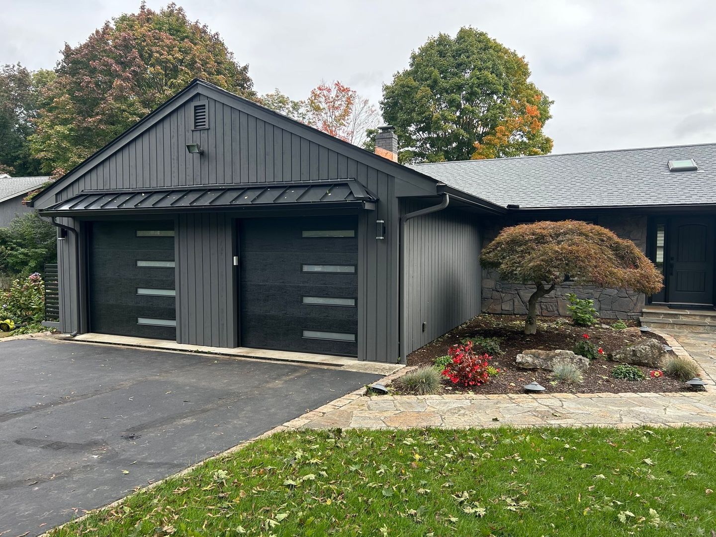 Dark gray two-car garage with driveway, landscaped front yard, and trees in fall colors.