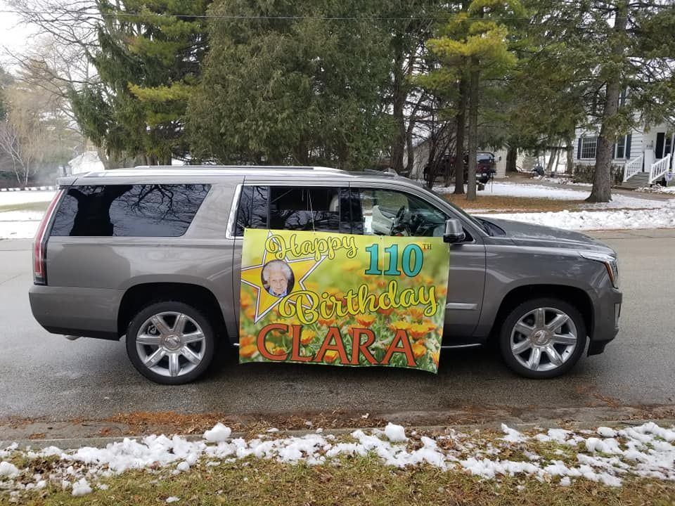 Gray SUV with banner Happy 110th Birthday CLARA parked on a snow-lined street.