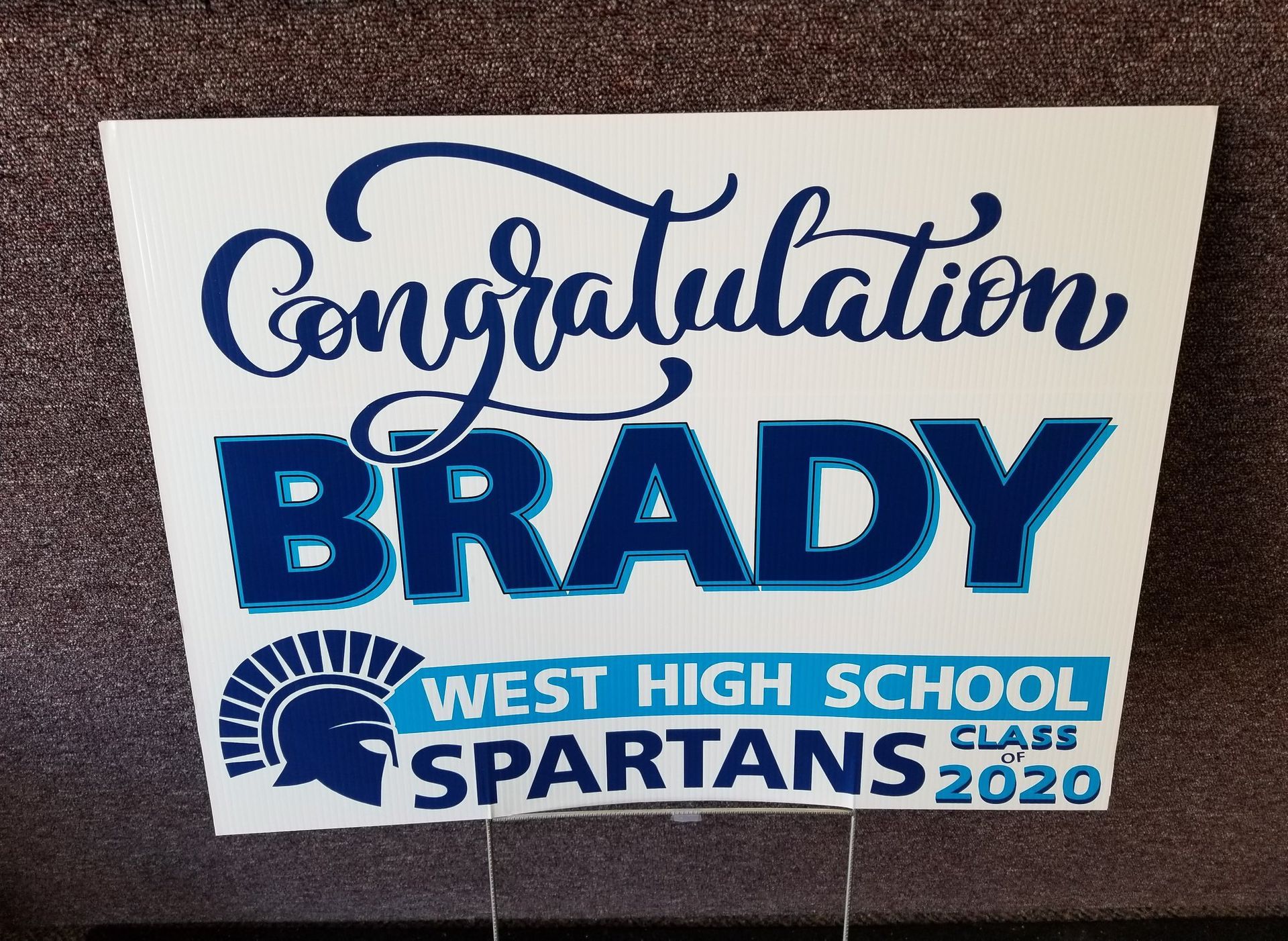 White graduation yard sign, blue text, Congratulations BRADY, West High School Spartans, Class 2020, with a Spartan helmet graphic.