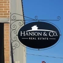 Hanson & Co. Real Estate sign, black oval, white text, hanging from ornate metal bracket, against blue sky.