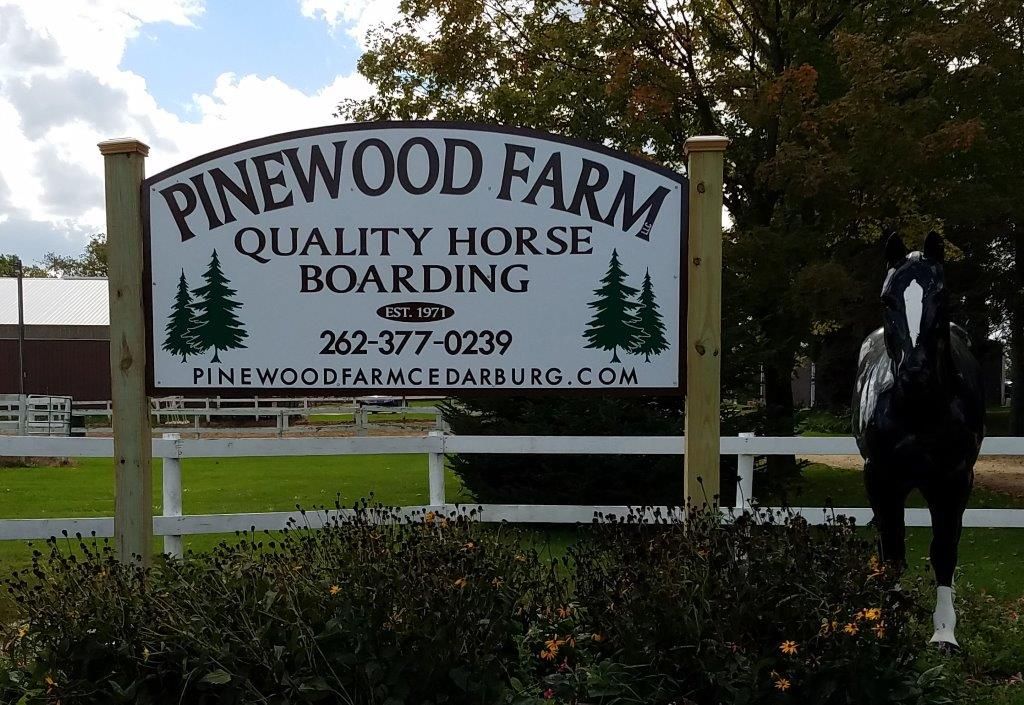Pinewood Farm sign with a horse near a white fence on a sunny day.
