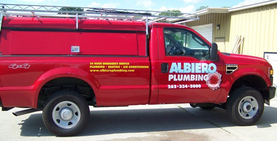 Red Albiero Plumbing truck parked outside a building, with an equipment rack on the roof.